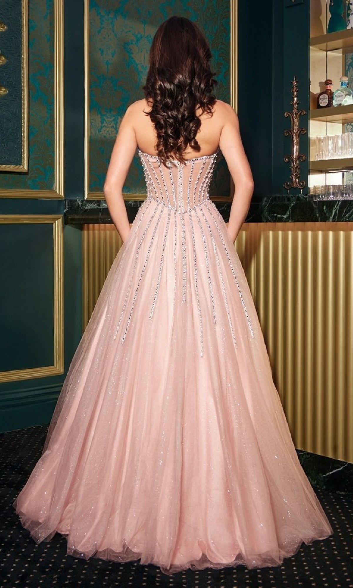 A woman with long brown hair models the Ladivine CDS541, a strapless beaded pink prom ball gown, its fitted bodice sparkling as she poses with her back to the camera in an ornate room.